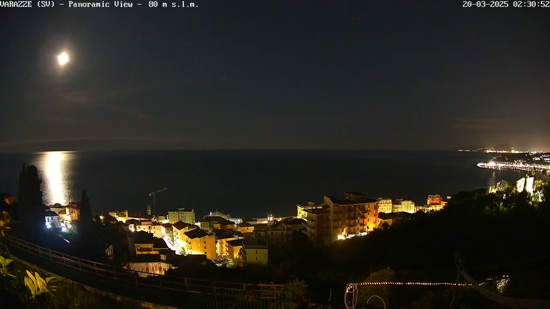 A nighttime panoramic view shows a coastal town illuminated under a bright moon, with a calm sea extending to a dimly lit coastline in the distance.