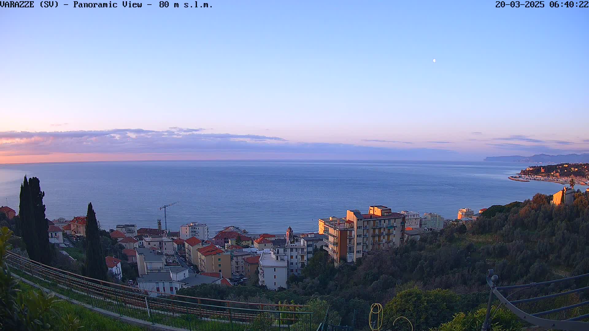 A panoramic view shows a calm sea at sunset, with a crescent moon in the sky, overlooking a coastal town nestled among green hills.