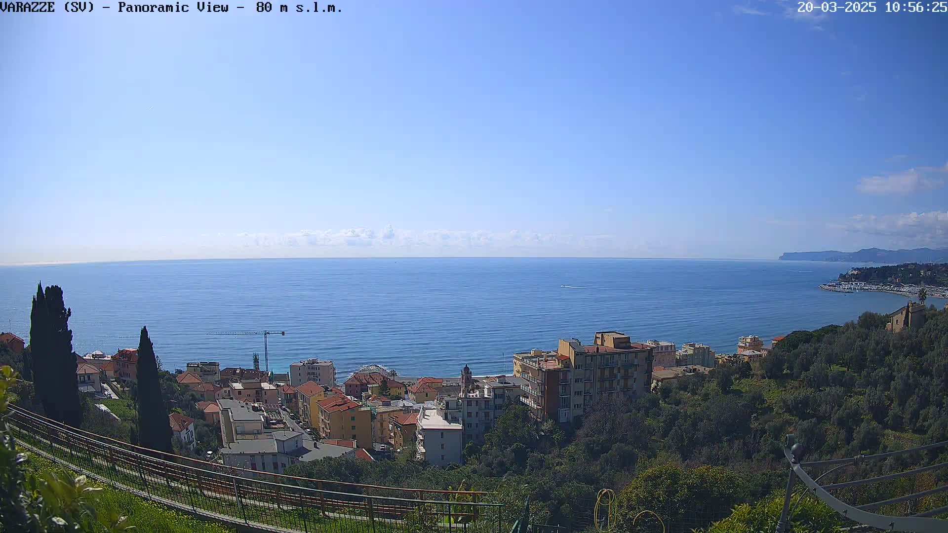 A coastal town nestled on a hillside overlooks a calm, blue sea under a mostly clear sky.