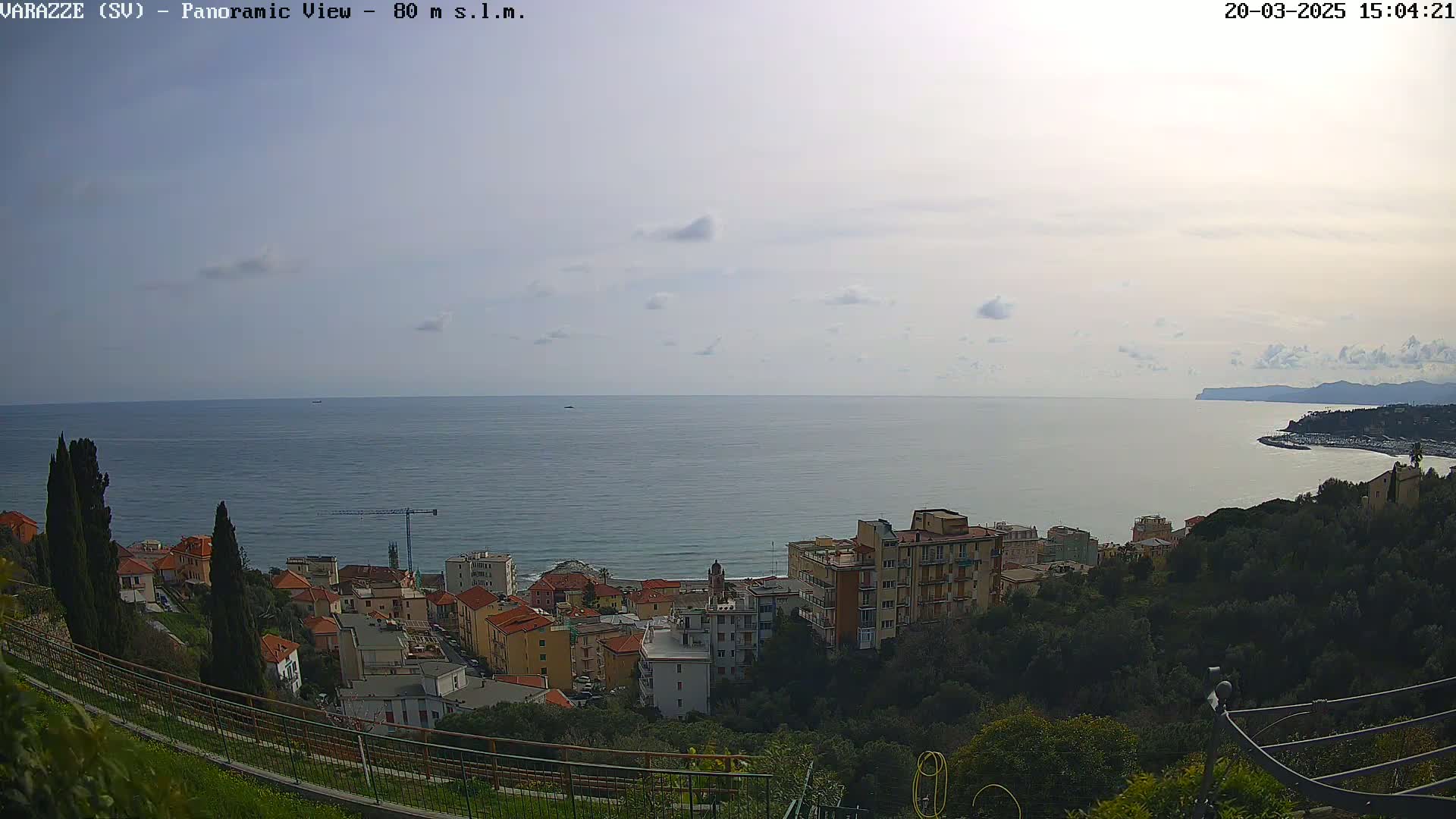 A coastal town nestled among green hills overlooks a calm sea under a partly cloudy sky.
