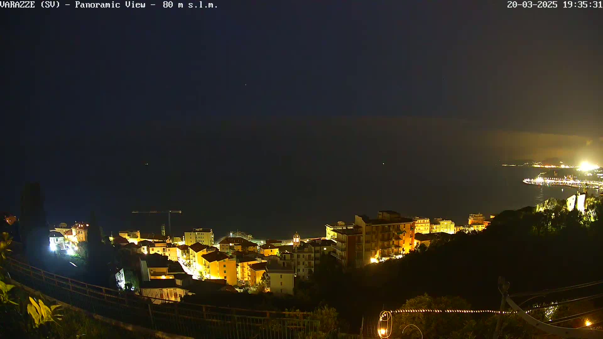 A nighttime, panoramic view shows a coastal town with illuminated buildings, overlooking a dark body of water under a clear, dark sky.