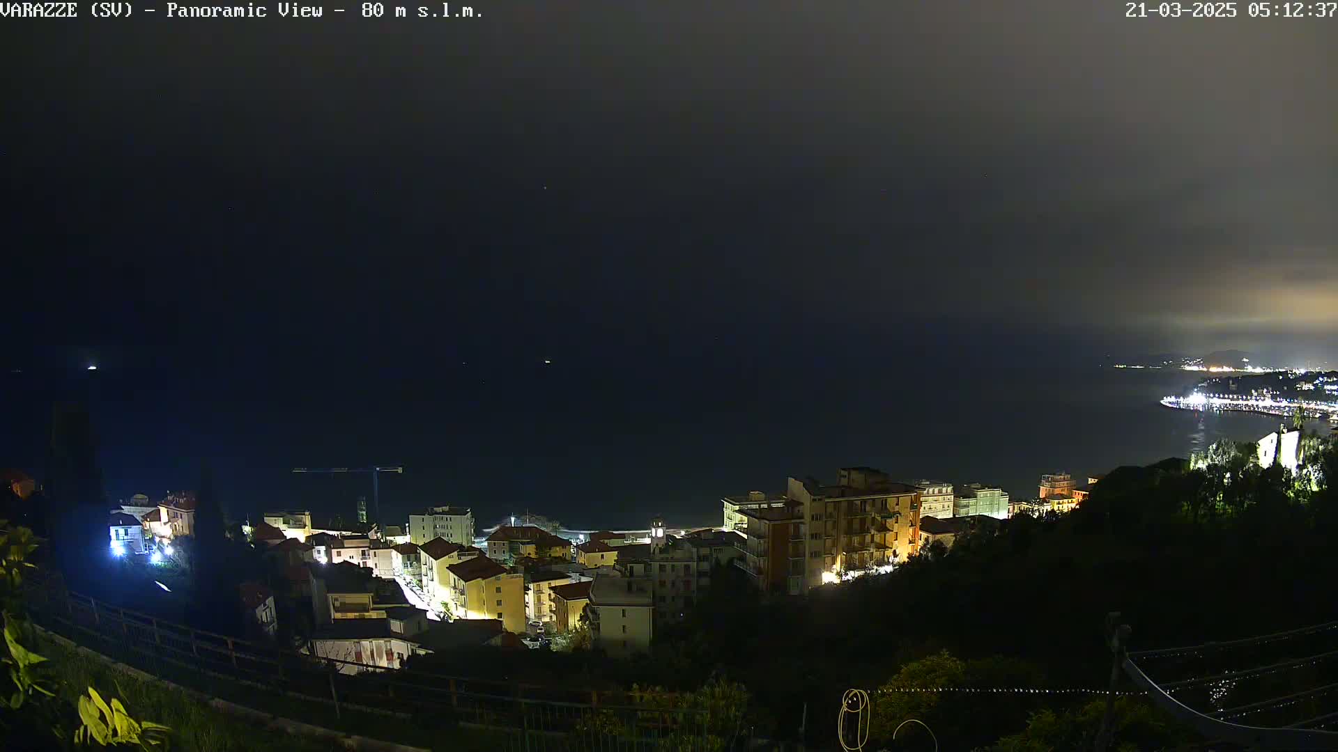 A nighttime panoramic view shows a coastal town illuminated, with a dark, calm sea extending to a dimly lit coastline in the distance under a mostly cloudy sky.