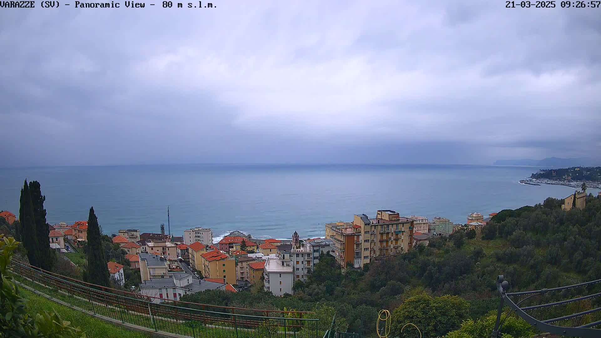 A coastal town with multi-story buildings is nestled on a hillside overlooking a calm sea under an overcast sky.