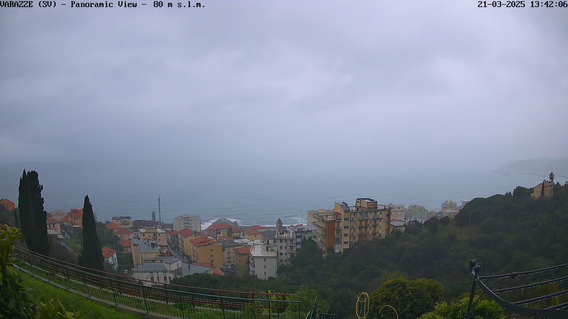 A coastal town is viewed from a hilltop on an overcast day, with a hazy ocean visible in the distance.