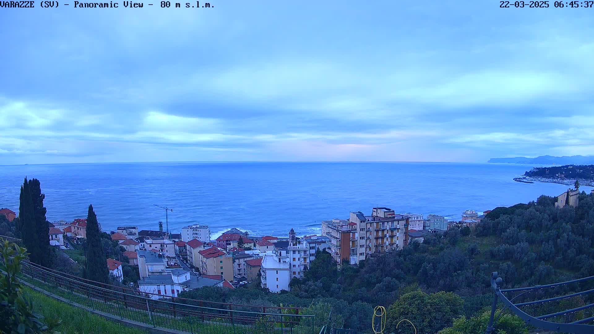 A coastal town nestled on a hillside overlooks a calm, blue sea under a mostly cloudy sky.