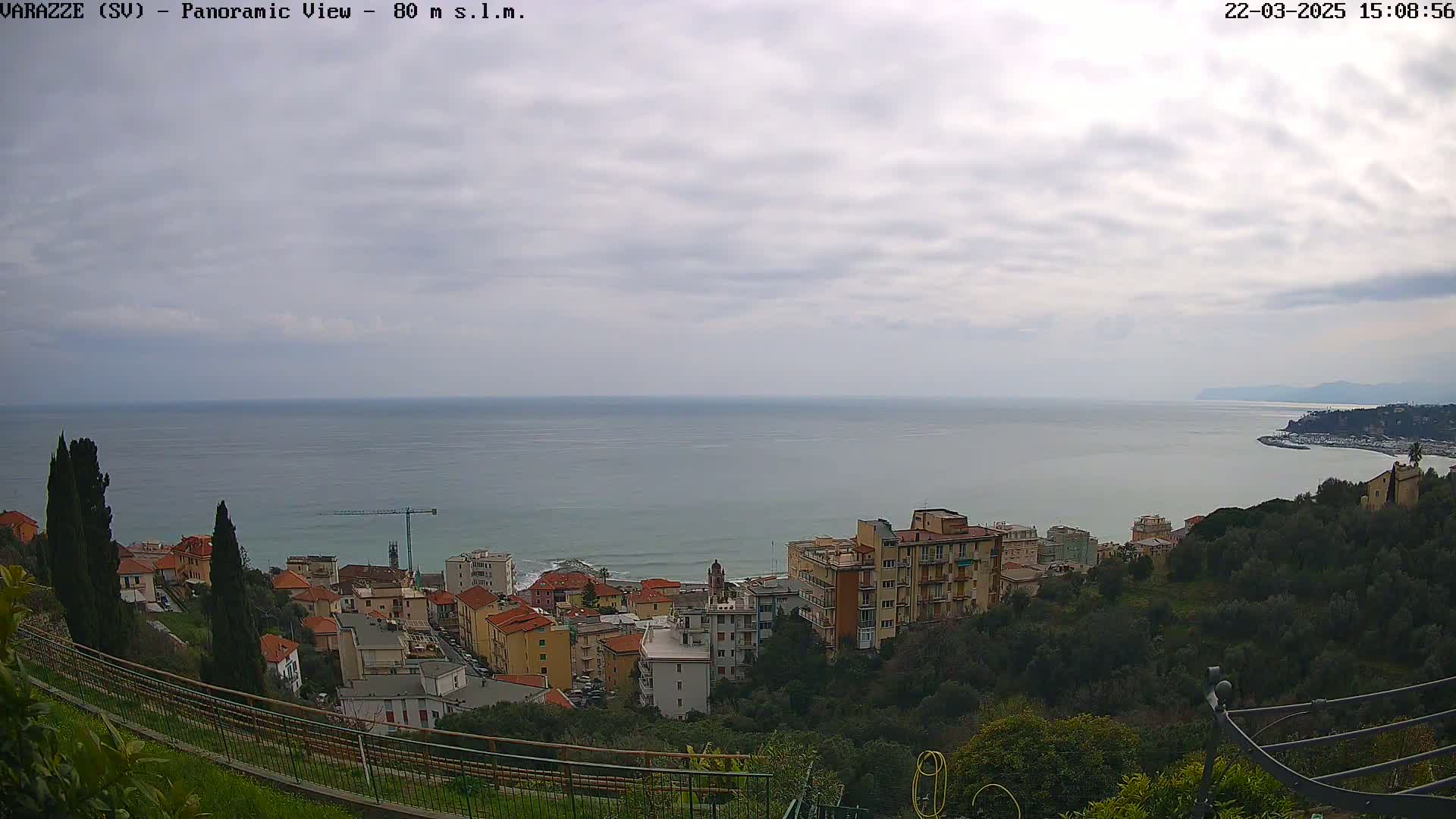 A panoramic view of a coastal town on a cloudy day shows a calm sea extending to the horizon, with buildings nestled on a hillside overlooking the water.