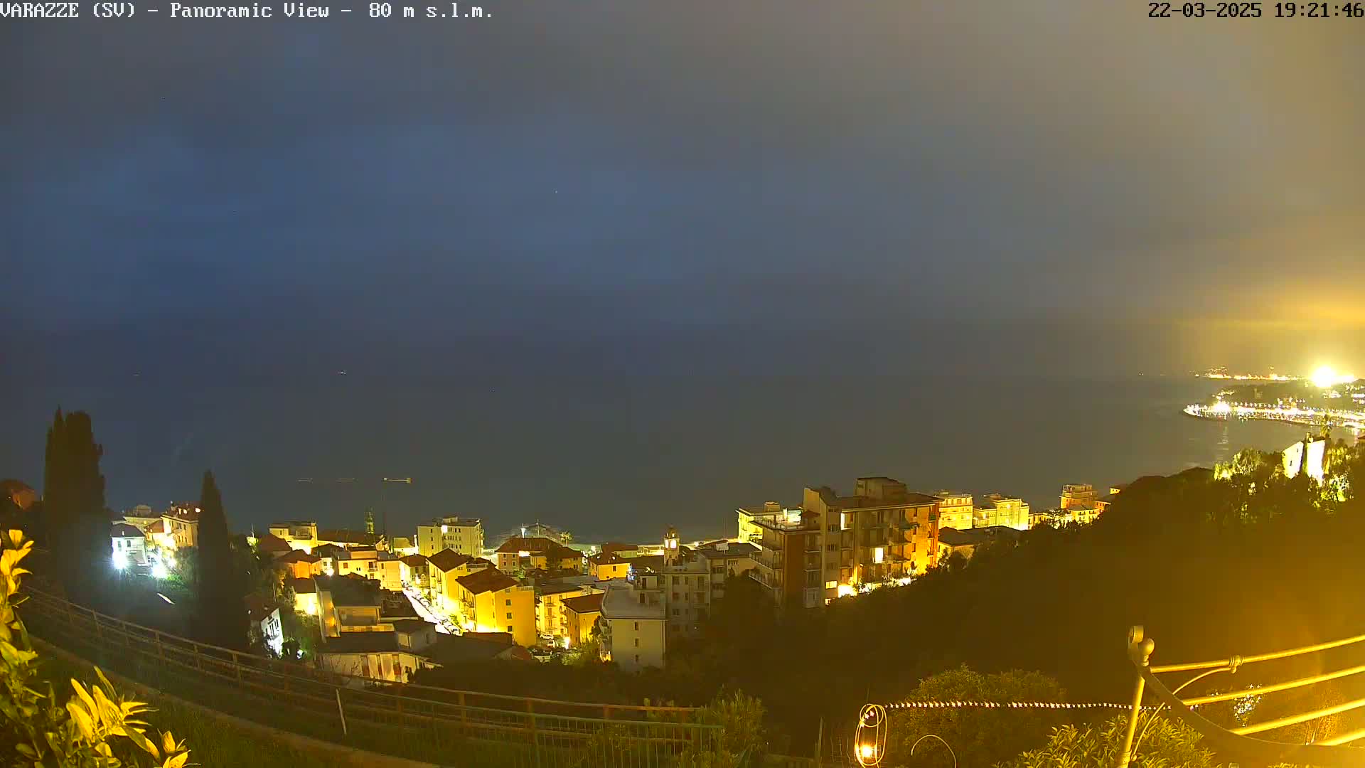 A nighttime panoramic view shows a coastal town illuminated with lights, overlooking a dark sea under a dusky sky.
