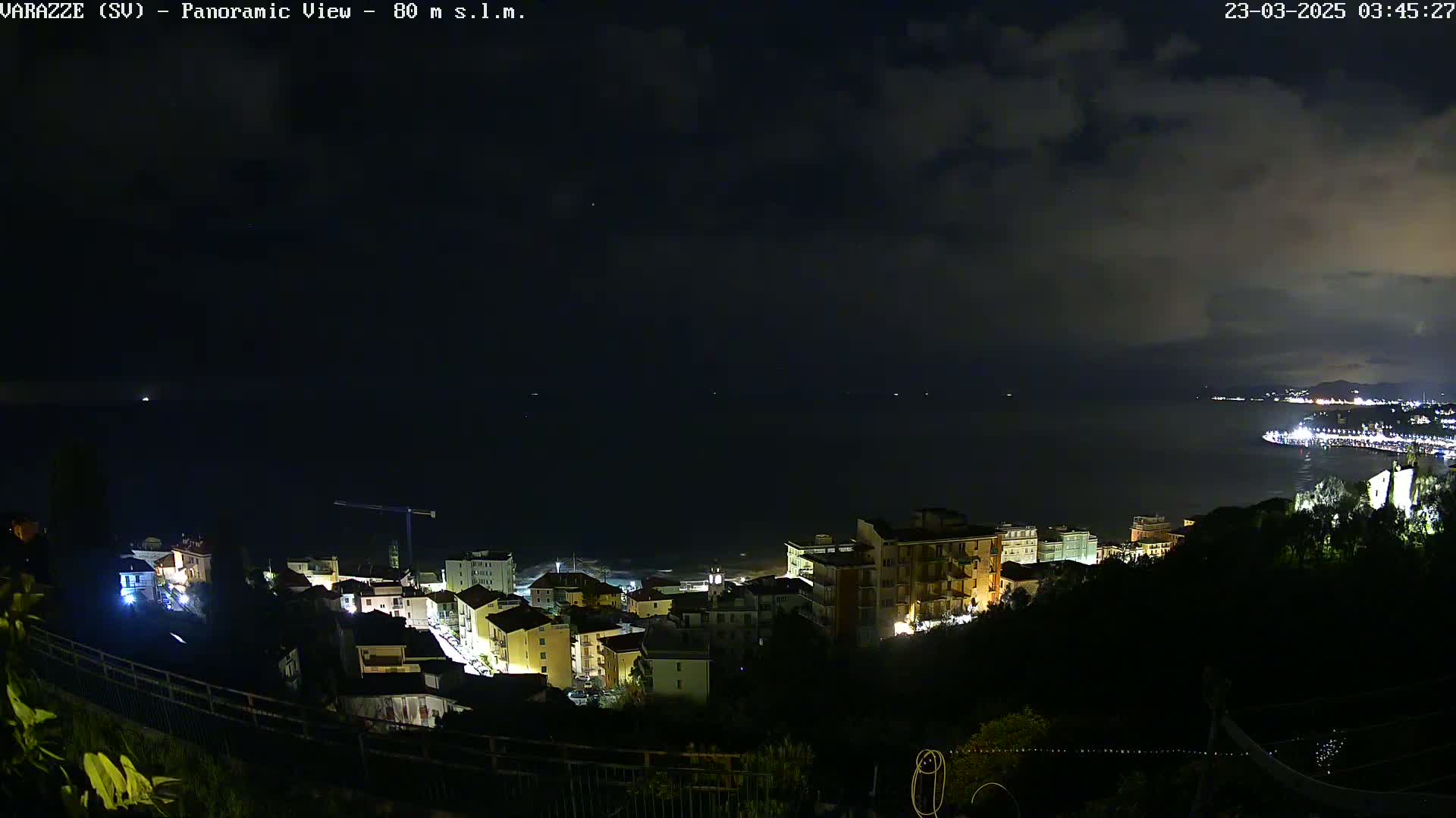 A nighttime panoramic view shows a coastal town with lights illuminating buildings and streets, overlooking a dark sea under a cloudy sky.
