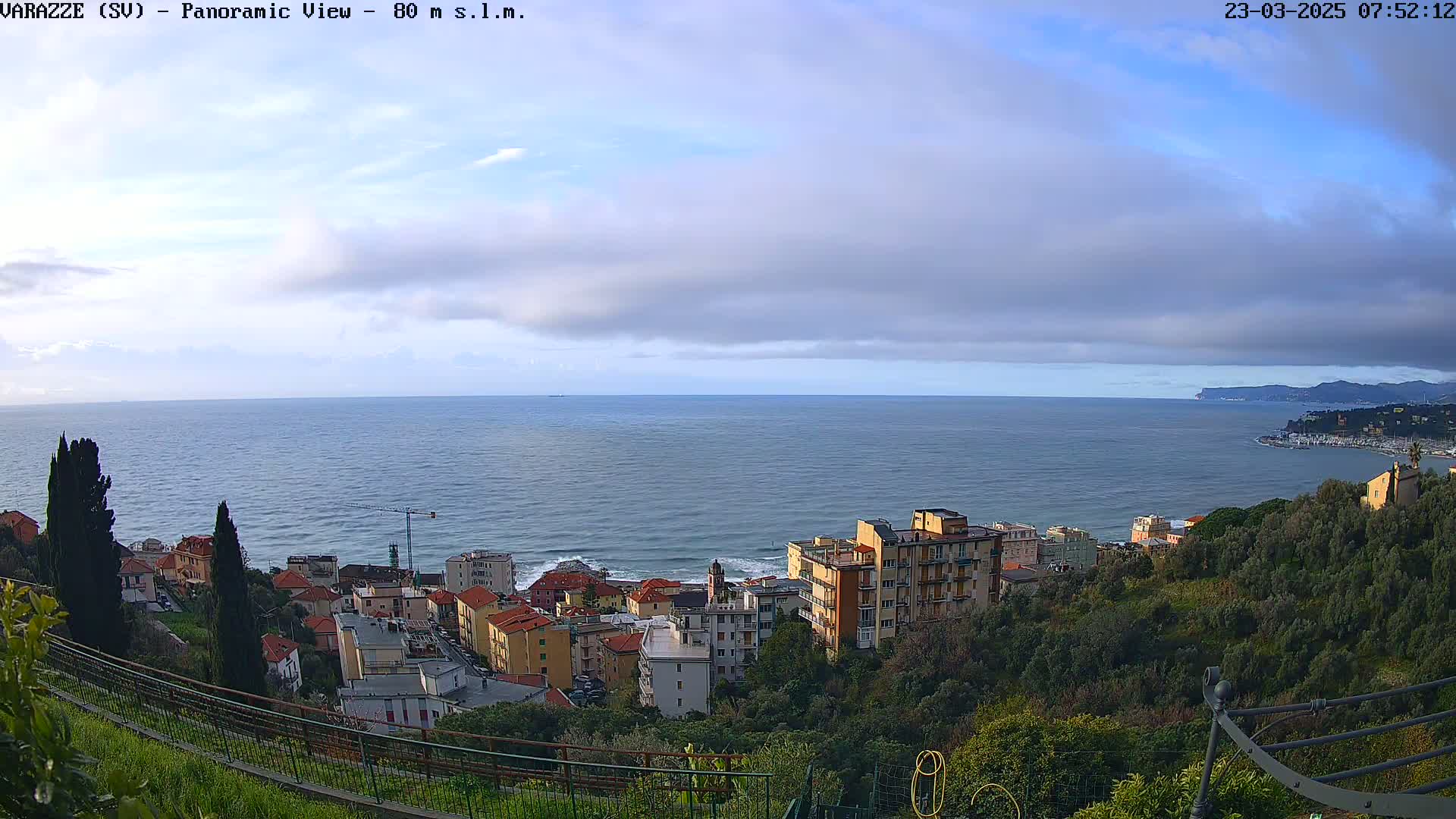 A coastal town with buildings nestled into a hillside overlooks a calm sea under a mostly cloudy sky.