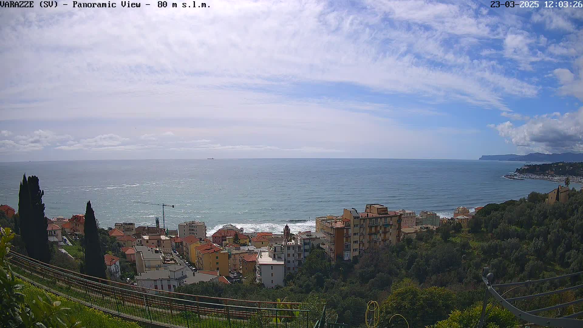 A panoramic view shows a coastal town nestled on a hillside overlooking a partly cloudy blue ocean, with some waves visible near the shore.