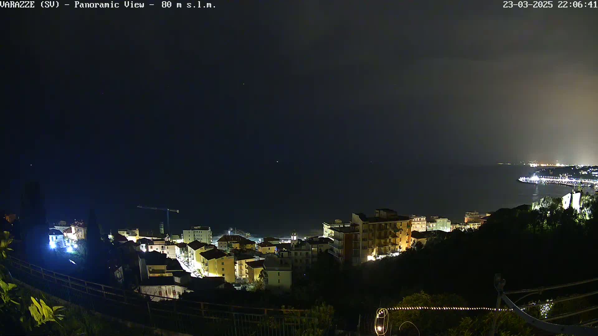 A nighttime panoramic view shows a coastal town illuminated, overlooking a dark sea under a mostly clear night sky.