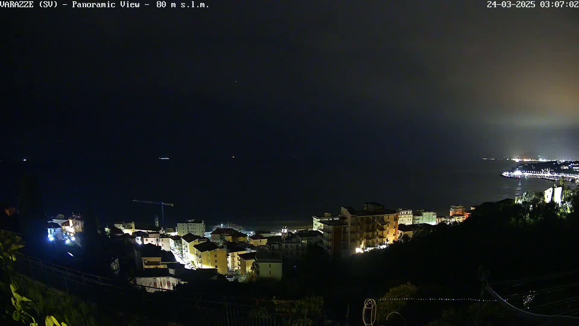 A nighttime panoramic view of a coastal town with dimly lit buildings and a dark, hazy sea.