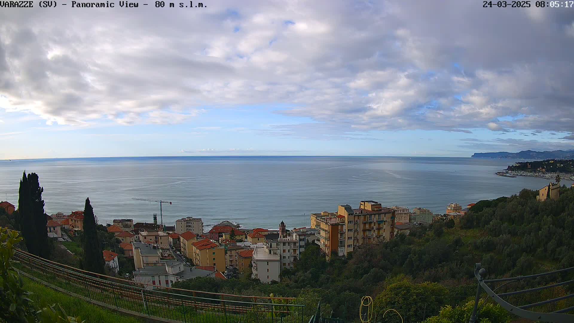 A panoramic view shows a coastal town nestled on a hillside overlooking a calm sea under a partly cloudy sky.