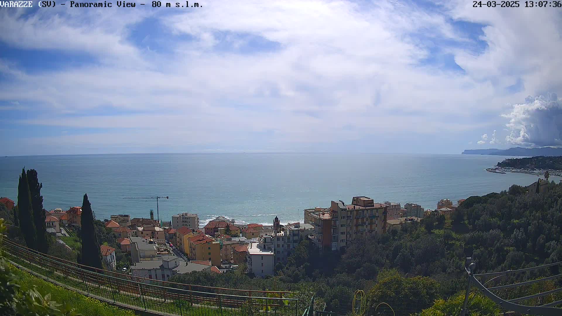 A partly cloudy sky overlooks a coastal town nestled on a hillside overlooking a calm sea.