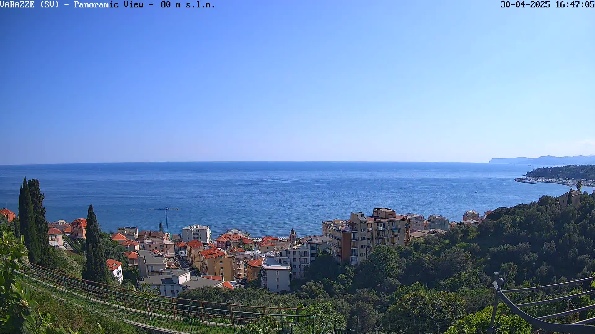 A coastal town with red-roofed buildings is nestled on a hillside overlooking a calm blue sea under a clear sky.