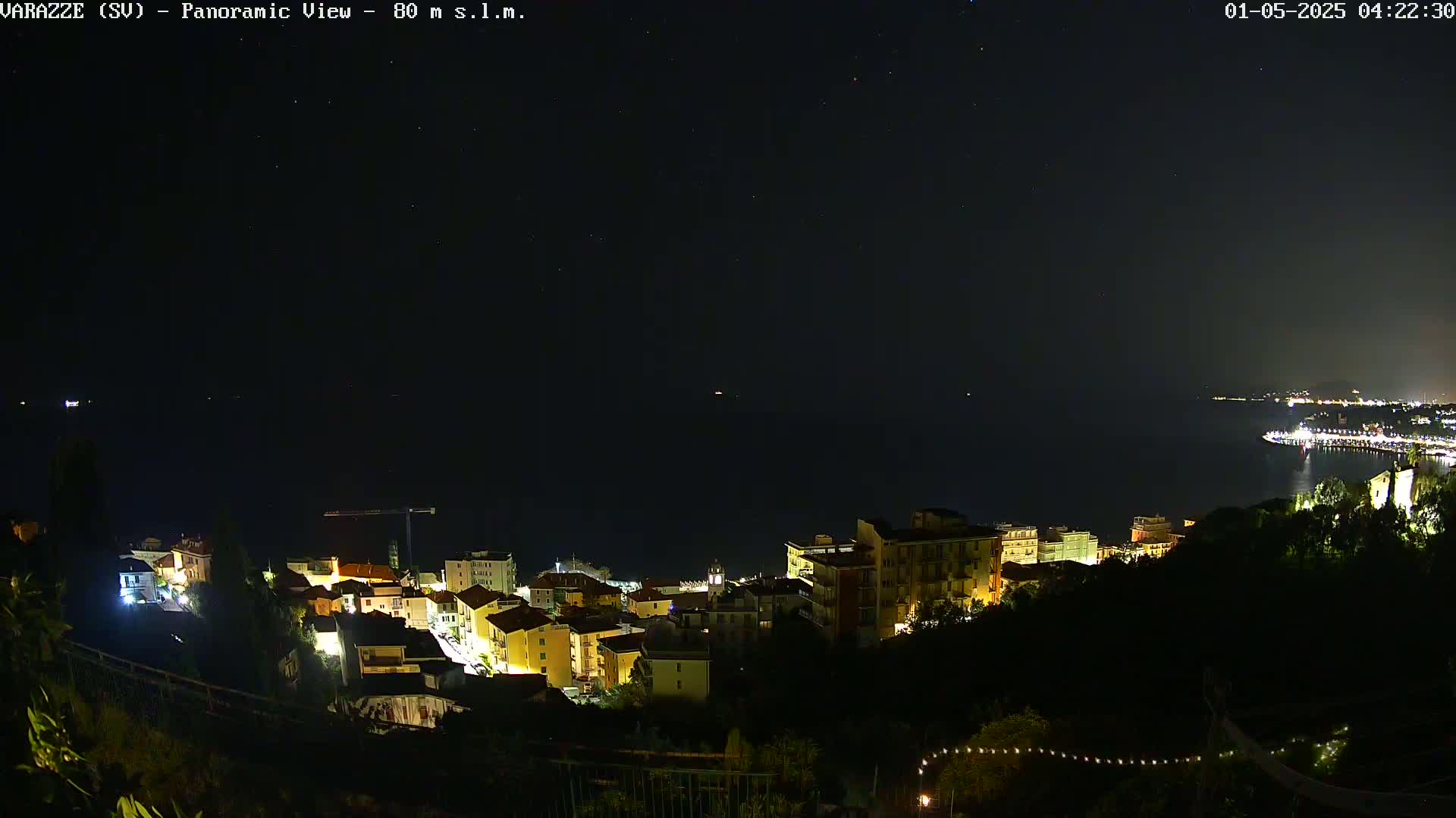A nighttime panoramic view shows a coastal town with its lights illuminated against a dark, clear sky dotted with stars.