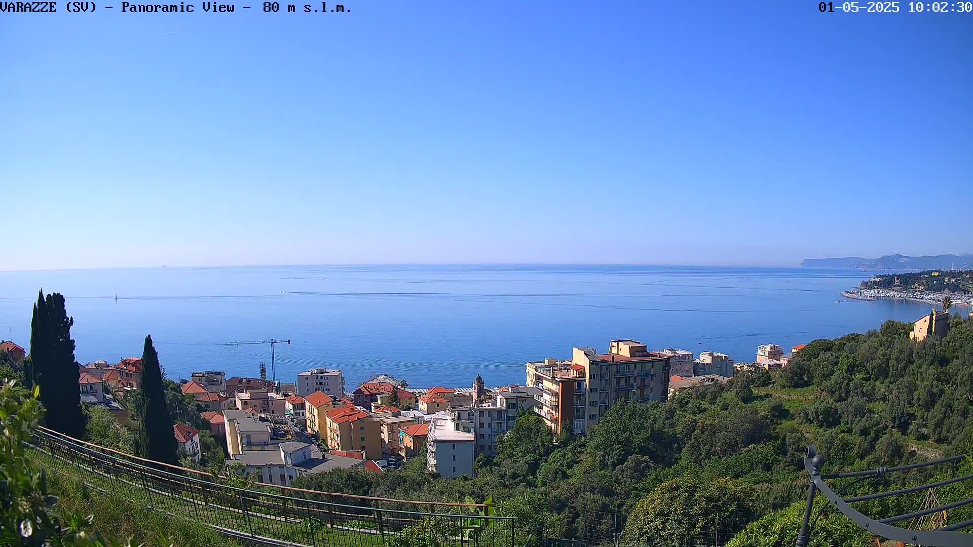A panoramic view shows a coastal town nestled amongst green hills overlooking a calm blue sea under a clear, bright sky.