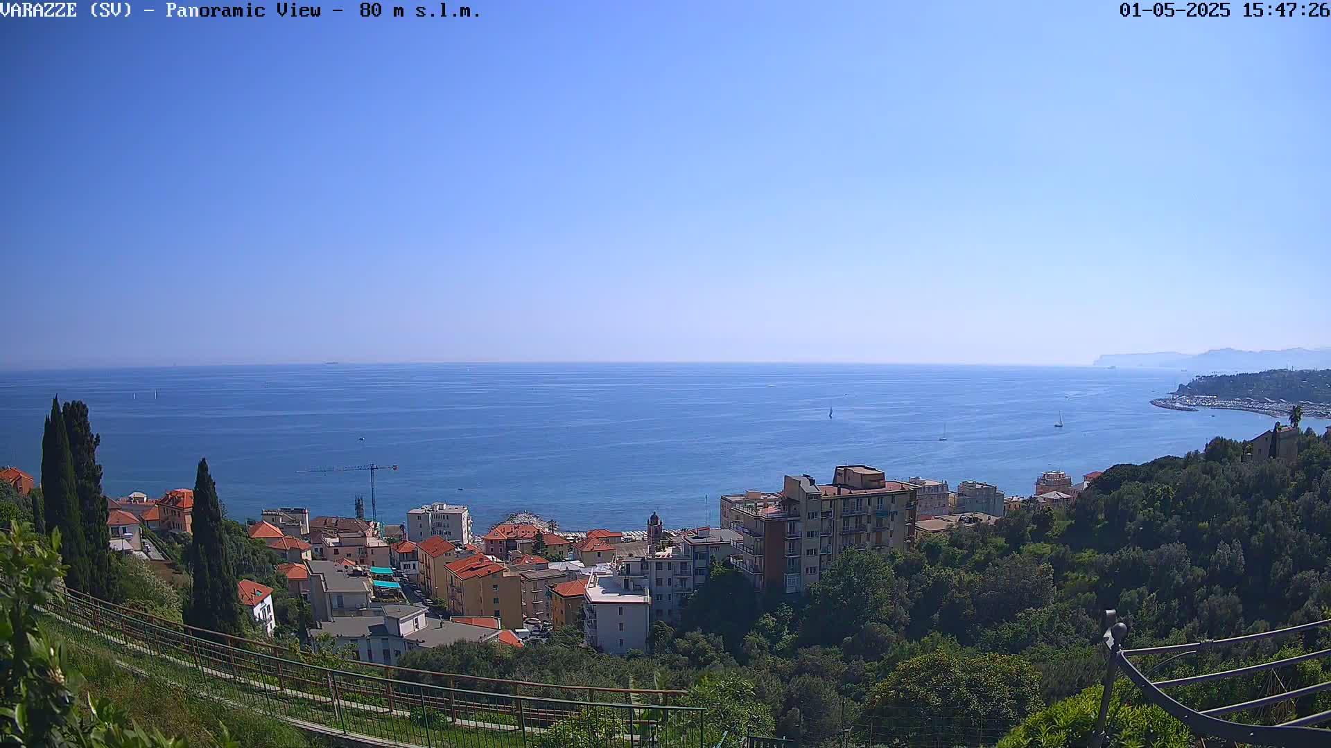 A panoramic view of a coastal town nestled on a hillside overlooking a calm, blue sea under a clear sky.