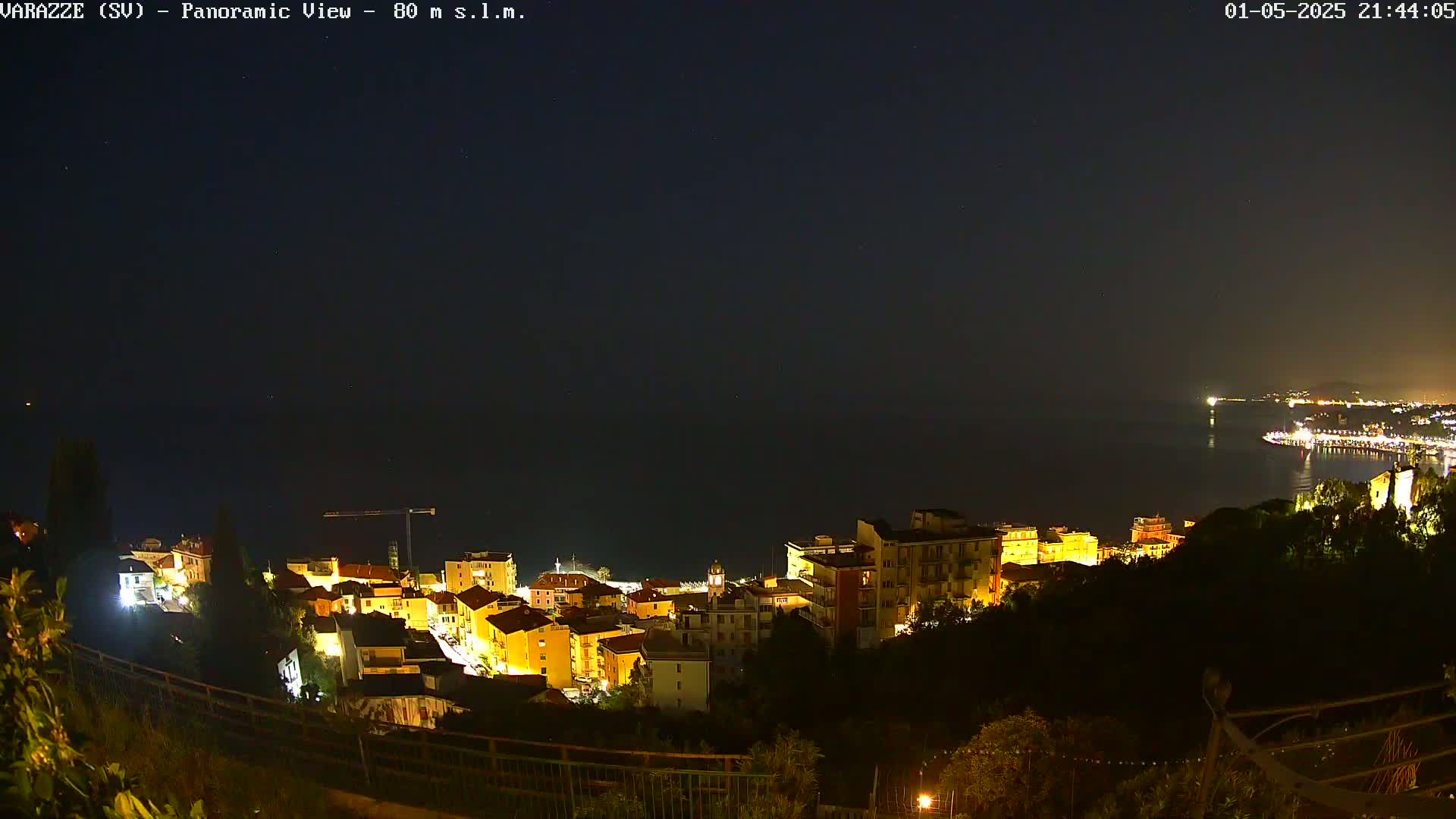 A nighttime panoramic view shows a coastal town illuminated, with a dark sea and clear night sky visible in the background.