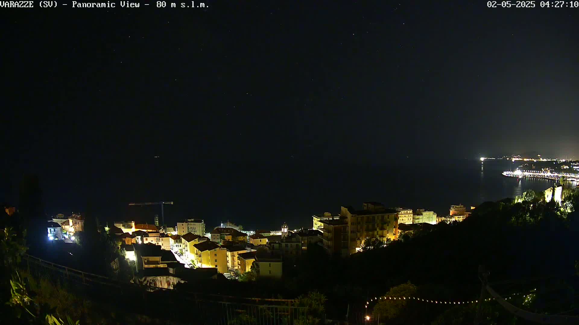 A nighttime, panoramic view shows a coastal town illuminated against a dark, starlit sky over calm water.