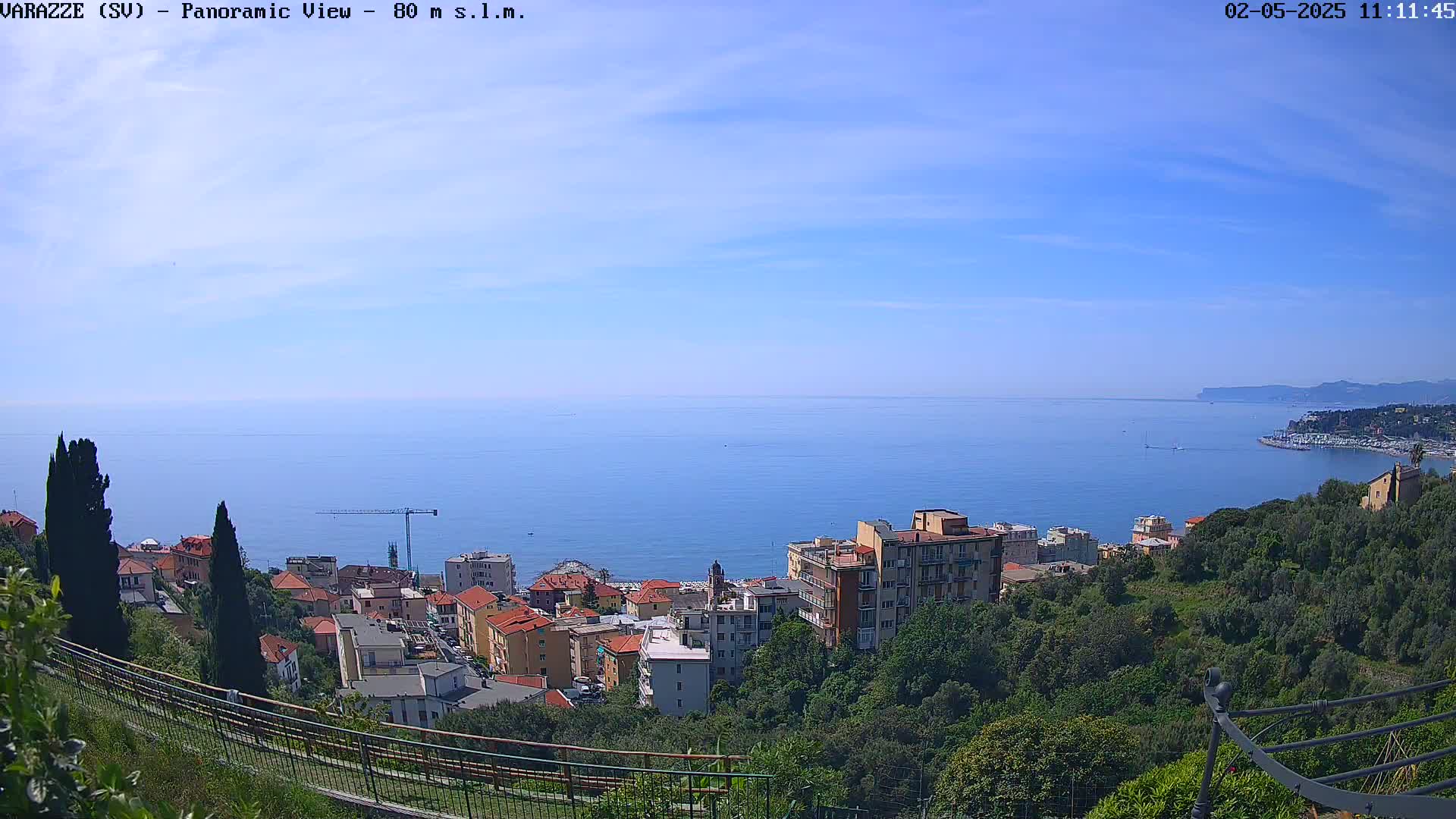 A panoramic view shows a coastal town nestled among green hills overlooking a calm blue sea under a clear, bright sky.