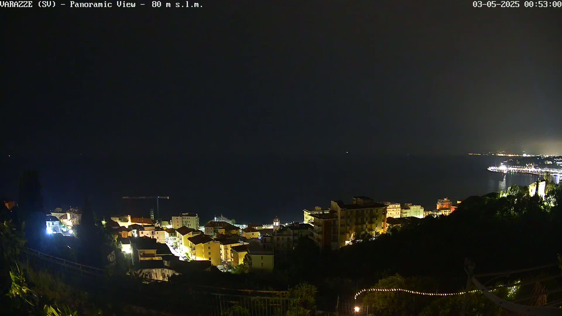 A nighttime panoramic view shows a coastal town illuminated with lights, overlooking a dark sea under a clear night sky.