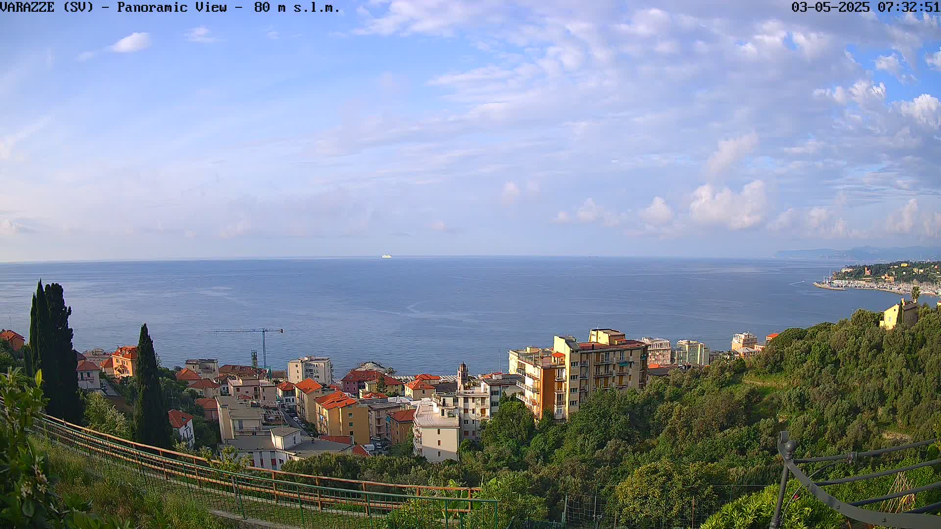 A panoramic view shows a coastal town nestled among green hills overlooking a calm sea under a partly cloudy sky.