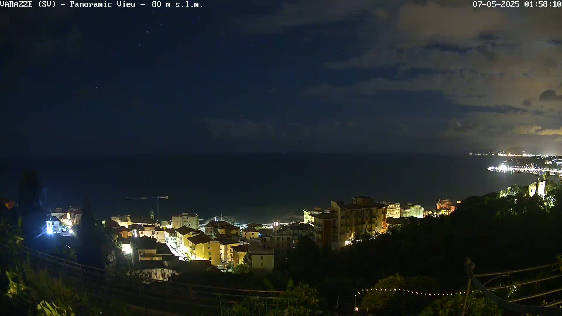 A nighttime panoramic view shows a coastal town illuminated, nestled on a hillside overlooking a dark sea under a partly cloudy sky.