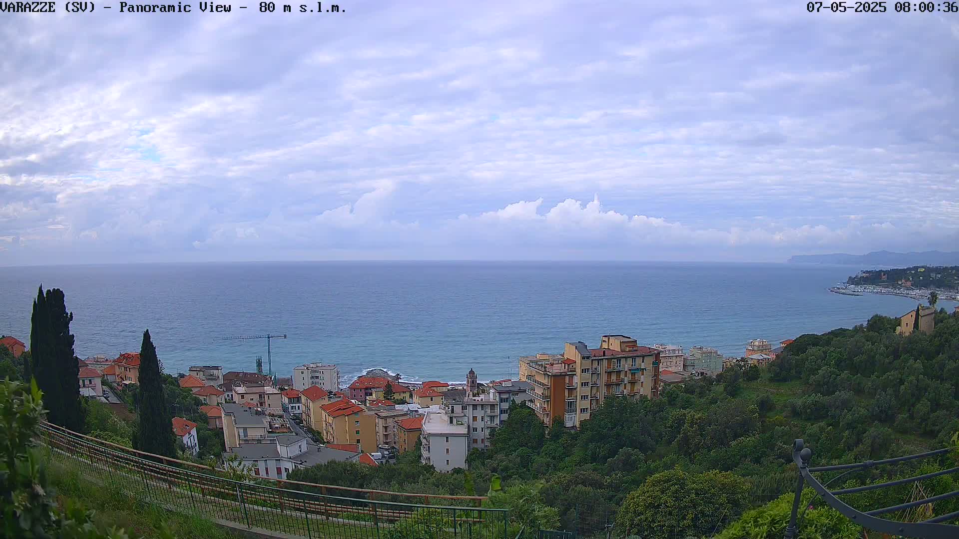 A coastal town with multi-story buildings sits on a hillside overlooking a calm blue sea under a partly cloudy sky.