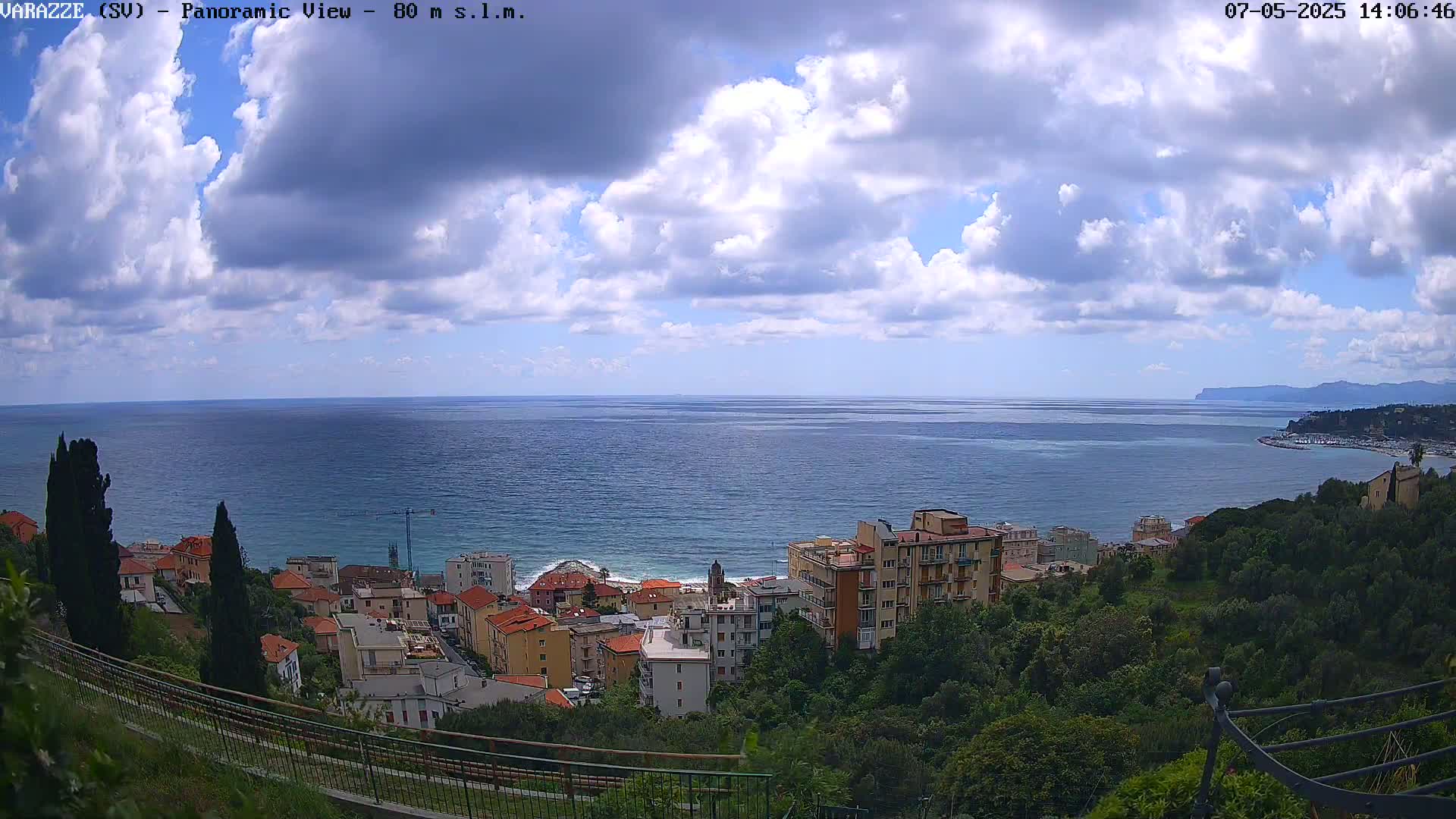 A partly cloudy day reveals a coastal town nestled on a hillside overlooking a calm sea, with distant mountains visible on the horizon.
