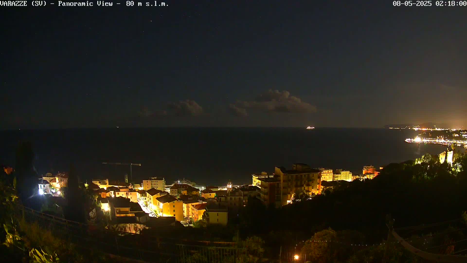 A nighttime, panoramic view of a coastal town with buildings illuminated, overlooking a dark sea under a partly cloudy sky.