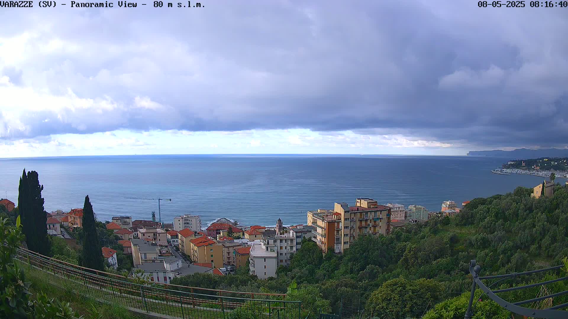 A coastal town with low buildings and green hills overlooks a calm sea under a partly cloudy sky.