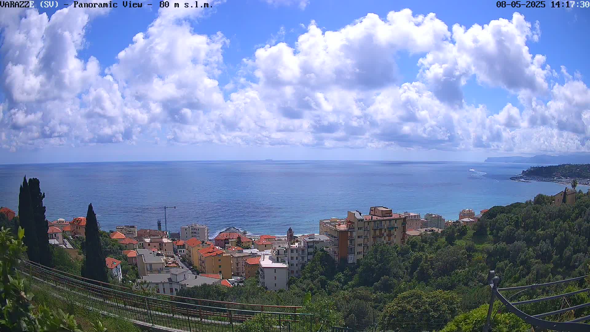 A panoramic view of a coastal town nestled amongst green hills, overlooking a calm blue sea under a partly cloudy sky.