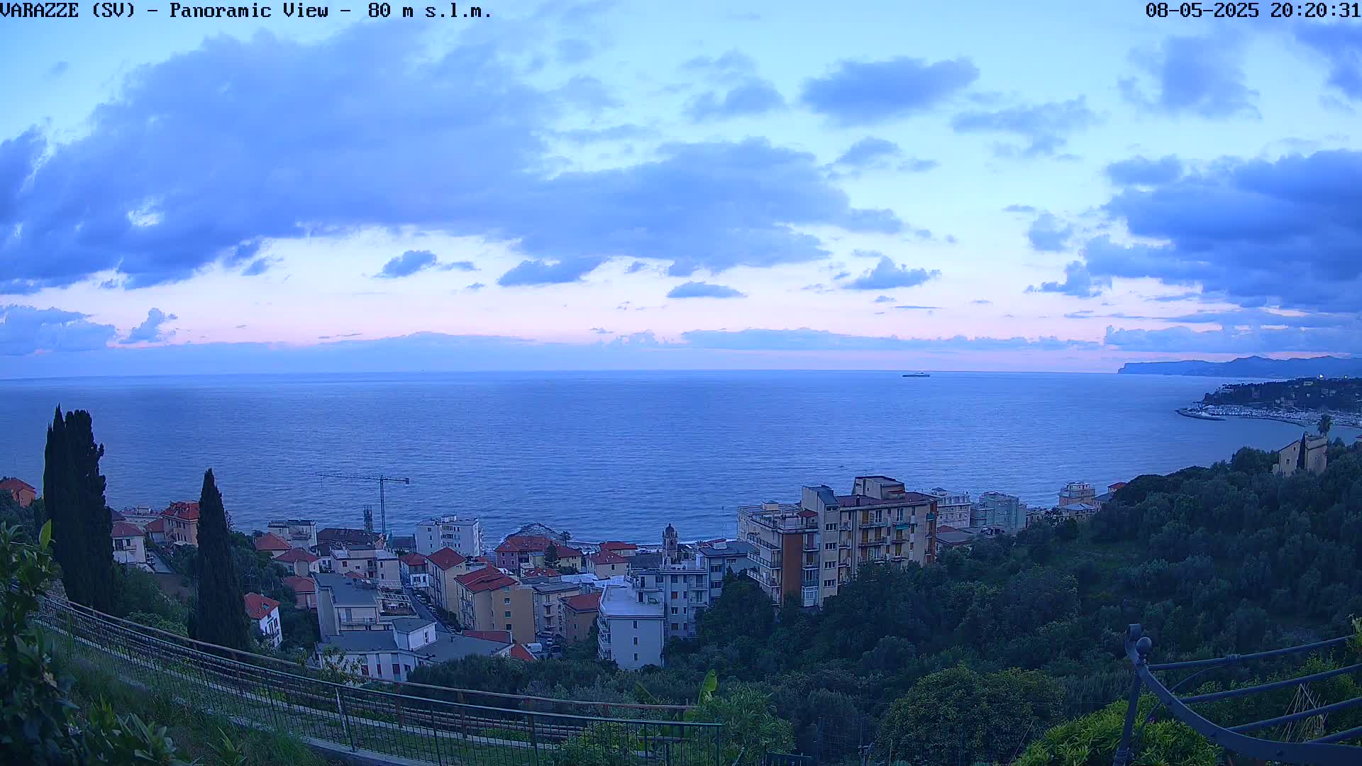 A coastal town nestled on a hillside overlooks a calm, twilight sea under a partly cloudy sky.