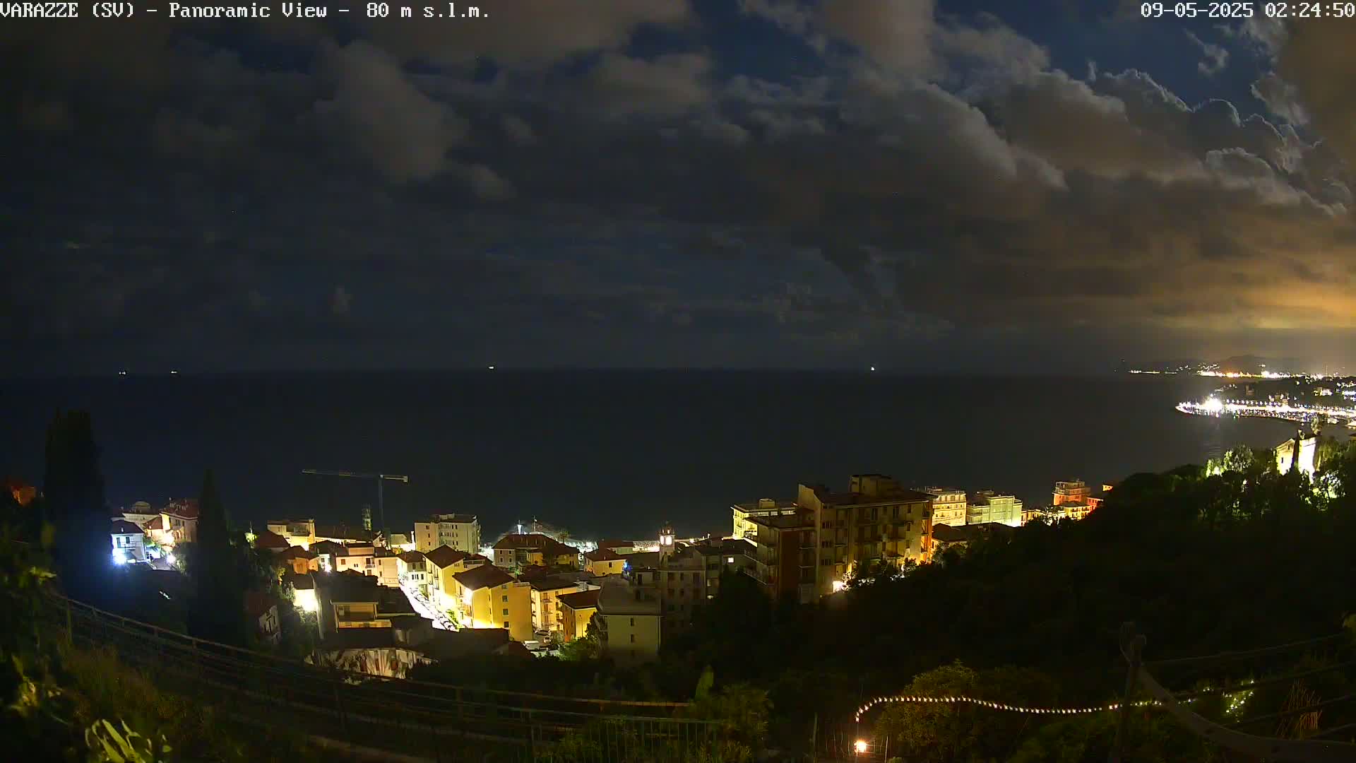 A nighttime panoramic view shows a coastal town with illuminated buildings nestled on a hillside overlooking a dark sea under a cloudy sky.
