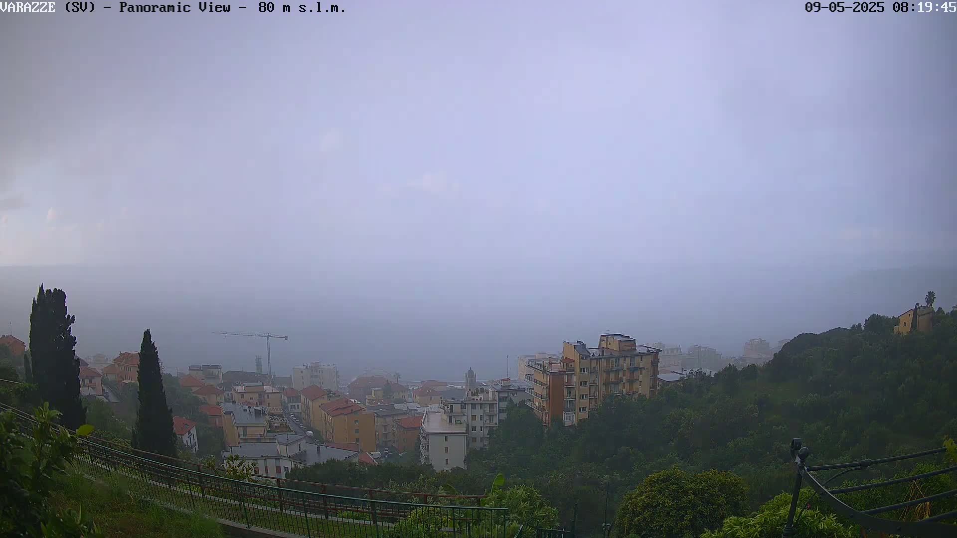 A hazy, rainy view overlooks a town nestled on a hillside overlooking the sea, with construction cranes visible in the distance.