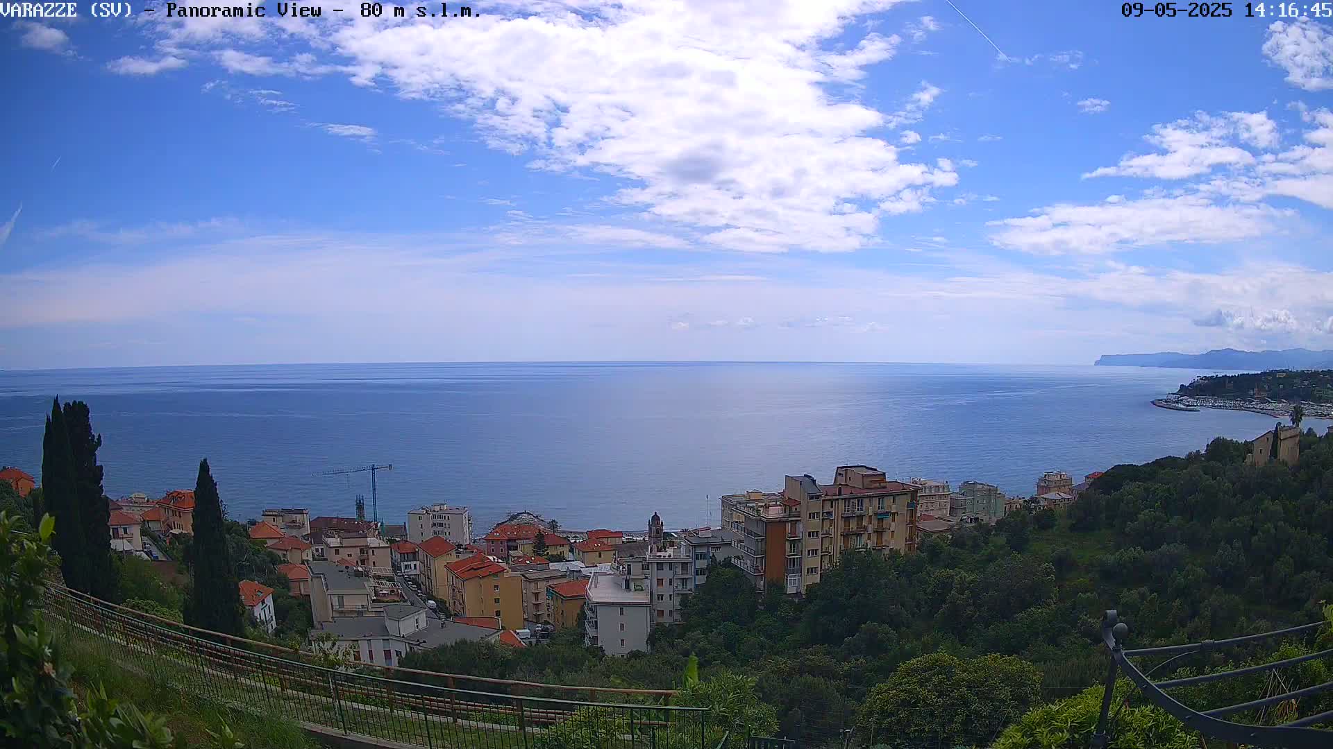 A coastal town nestled on a hillside overlooks a calm, blue sea under a partly cloudy sky.