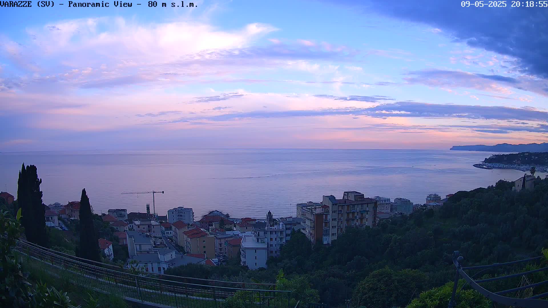 A panoramic view of a coastal town at dusk under a partly cloudy sky, showing a calm sea extending to a distant shoreline.