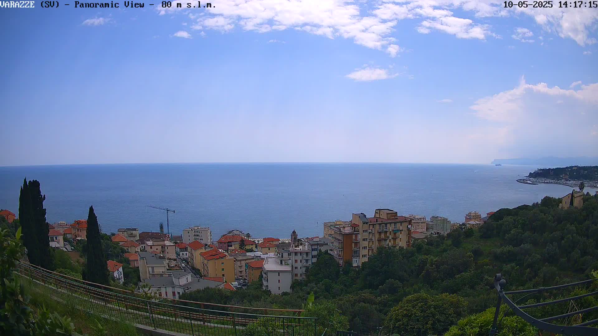 A panoramic view shows a partly cloudy sky over a calm blue sea, with a coastal town nestled among green hills in the foreground.