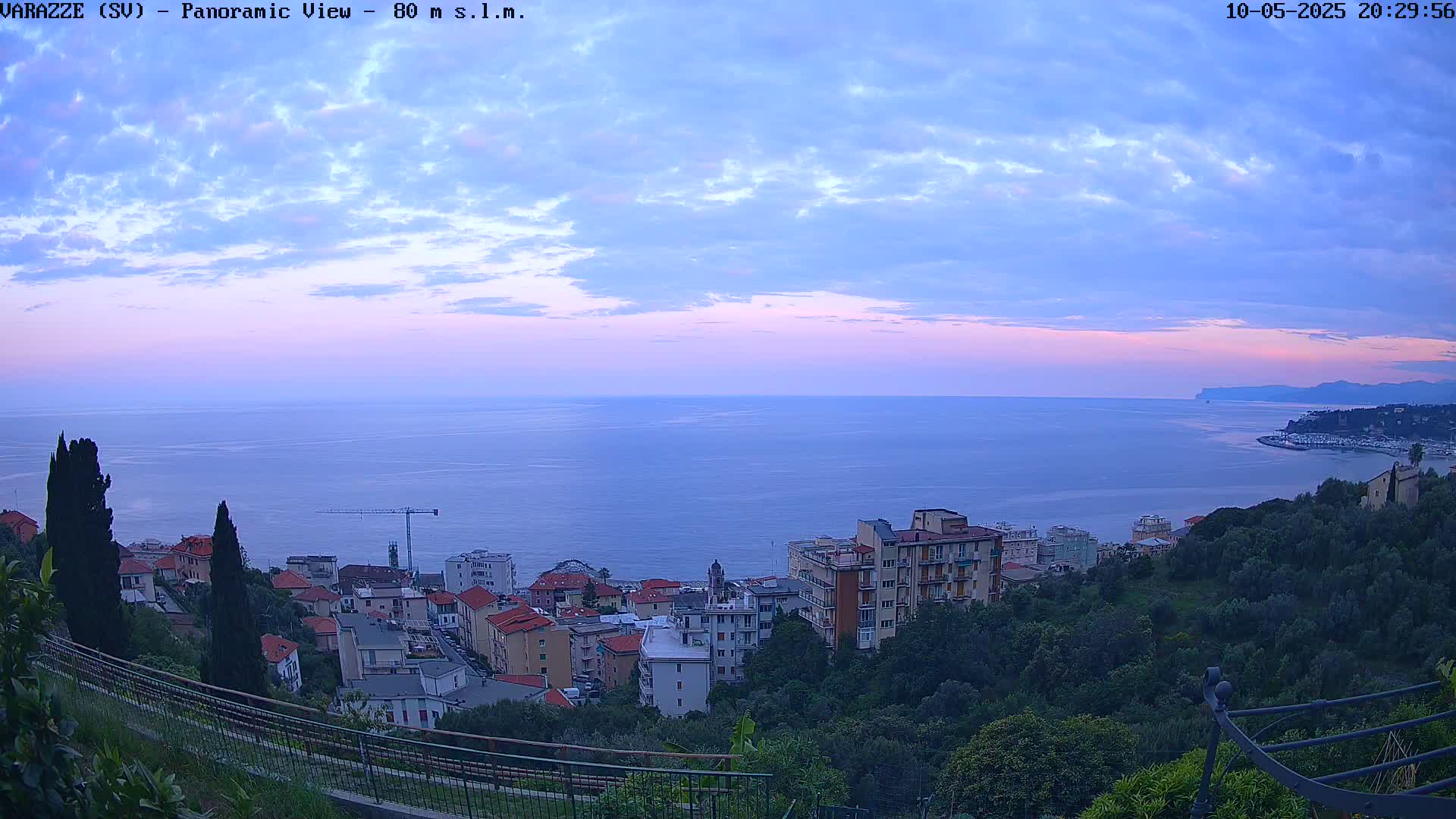 A panoramic view of a coastal town at dusk, under a partly cloudy sky, shows buildings nestled among green hills overlooking a calm sea extending to a distant shoreline.
