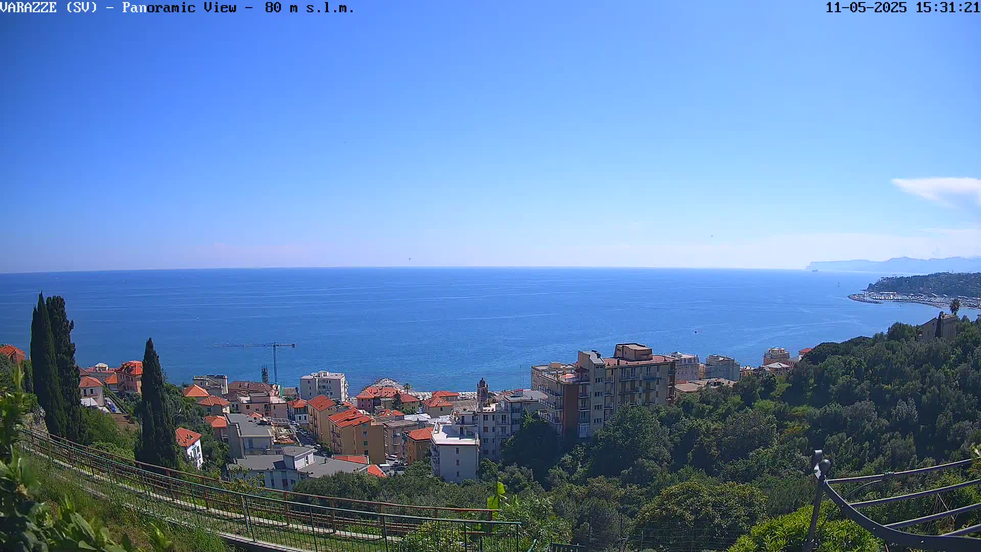A coastal town with low buildings nestled among green trees overlooks a calm, blue sea under a clear, sunny sky.