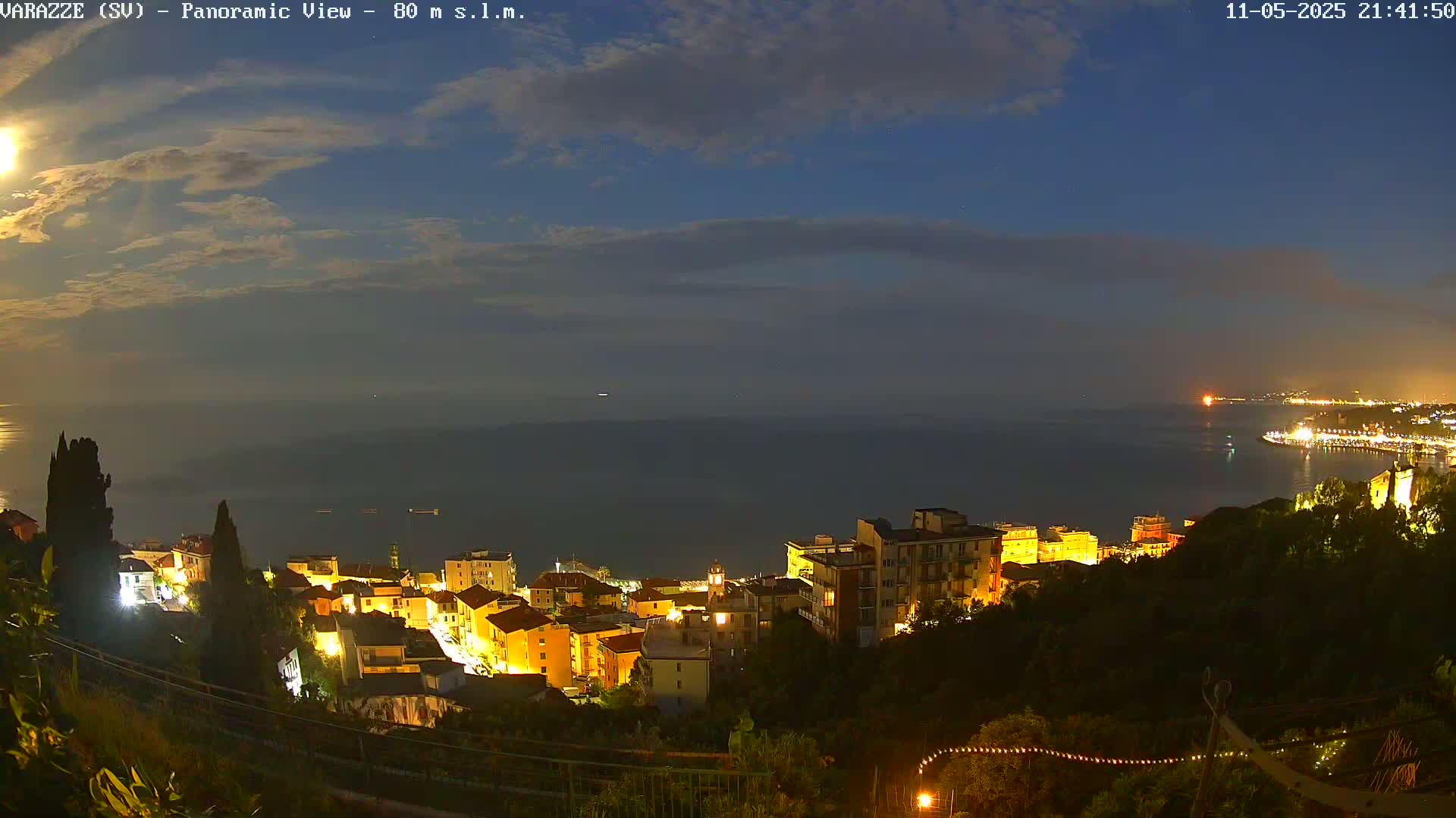 A nighttime panoramic view shows a coastal town illuminated, nestled on a hillside overlooking a calm sea under a partly cloudy sky with a bright moon.