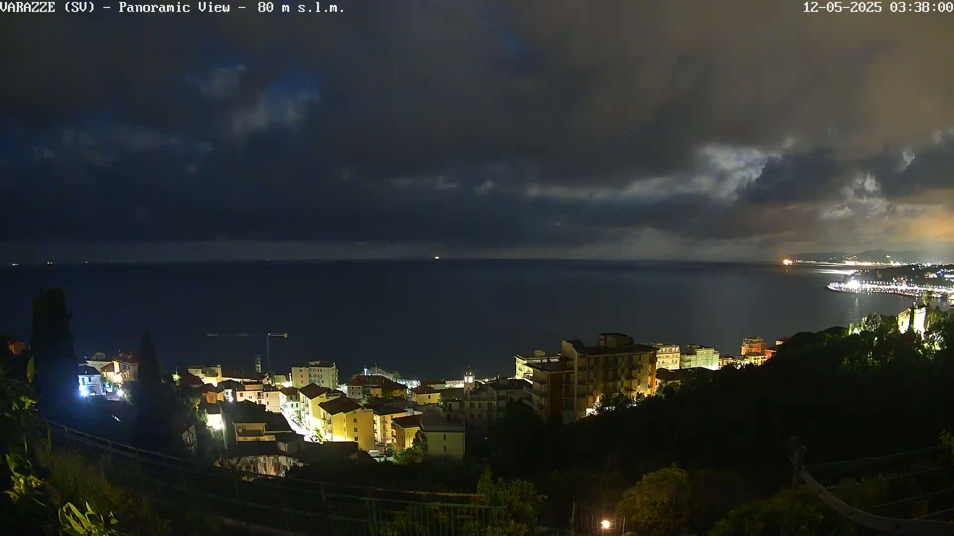 A nighttime view of a coastal town illuminated by streetlights, under a dark, cloudy sky with a calm sea extending to the horizon.