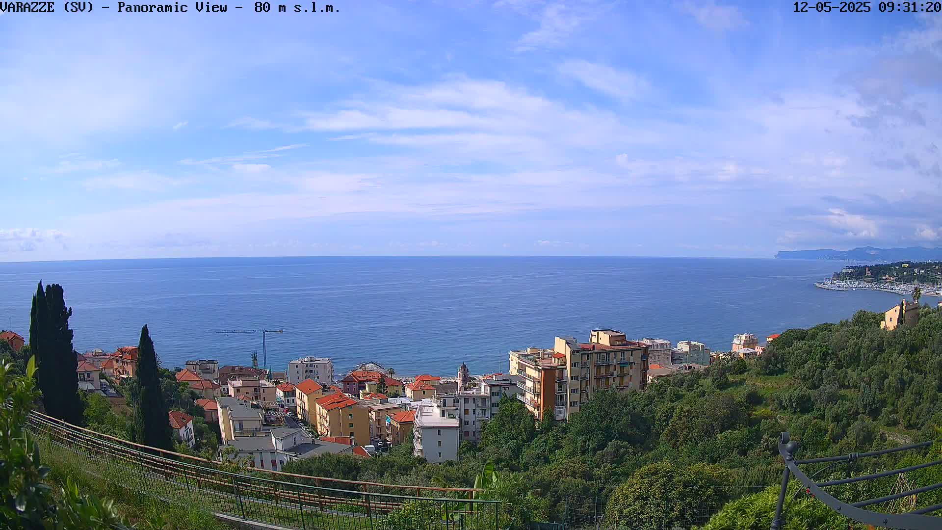 A panoramic view shows a mostly sunny day overlooking a coastal town nestled amongst lush green vegetation and a calm, blue ocean stretching to a distant shoreline.