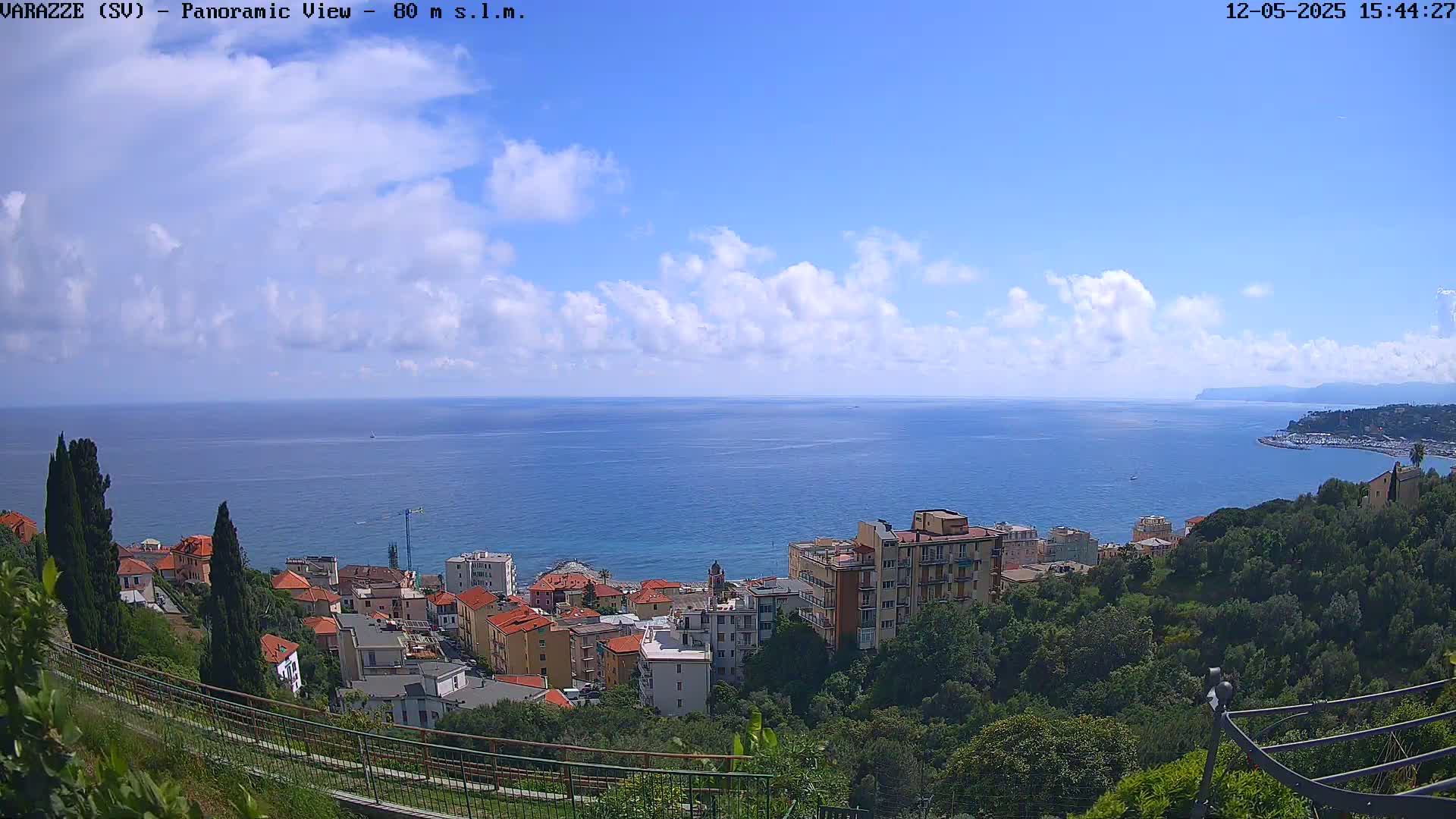 A coastal town nestled amongst green hills overlooks a calm blue sea under a mostly sunny sky with scattered clouds.