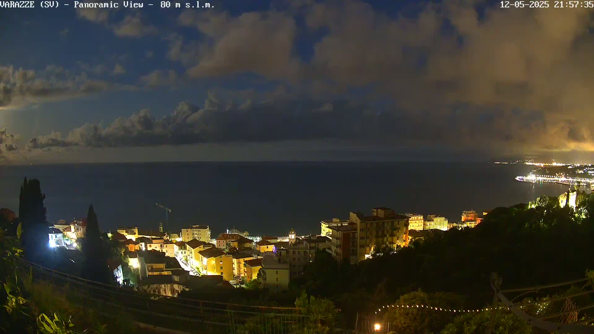 A nighttime panoramic view of a coastal town, illuminated with lights, under a partly cloudy sky, overlooking a dark sea.