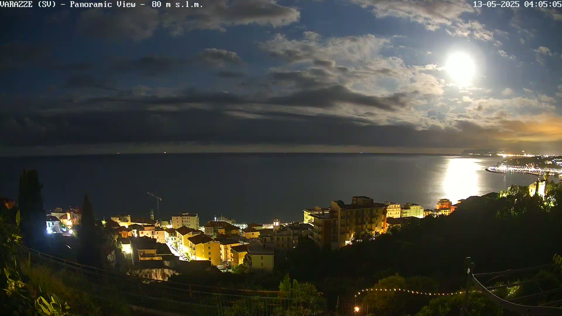 A nighttime panoramic view shows a coastal town illuminated under a partly cloudy sky with a bright moon.