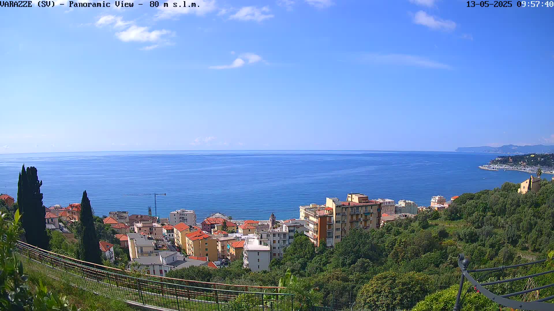 A coastal town with multi-story buildings is nestled among green hills overlooking a calm, blue sea under a clear, mostly sunny sky.