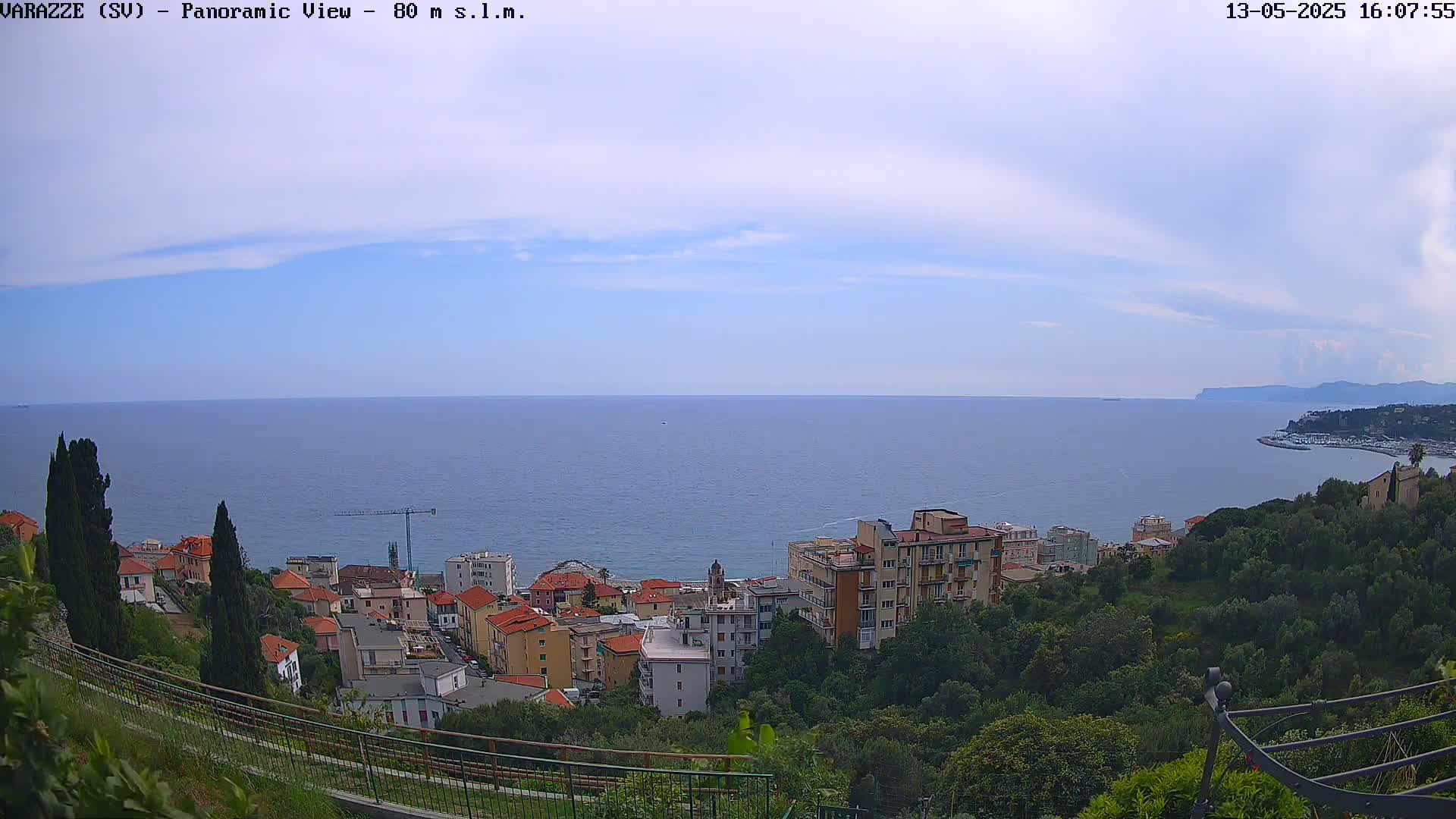A panoramic view of a coastal town shows a mostly clear sky and calm sea with buildings on a hillside overlooking the water.