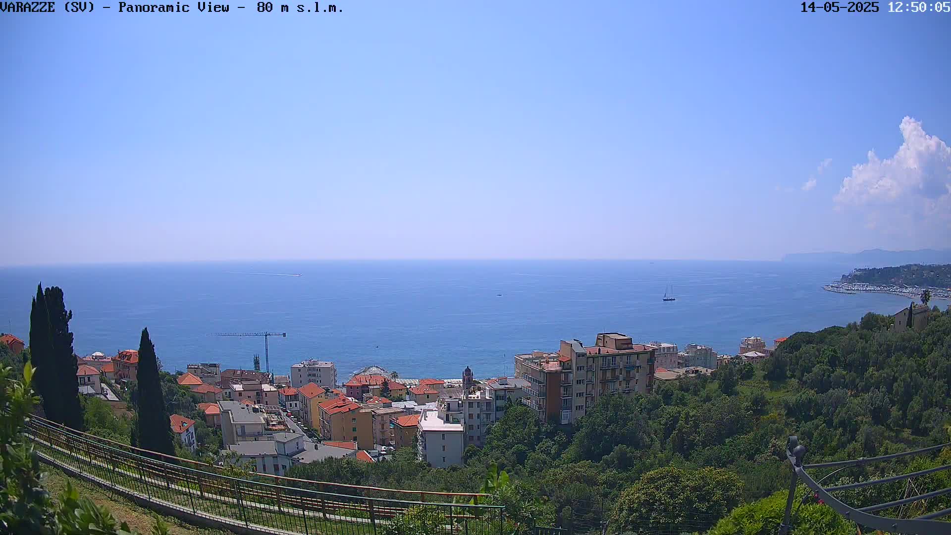A panoramic view shows a coastal town nestled amongst green hills overlooking a calm blue sea under a mostly clear sky with a few clouds.